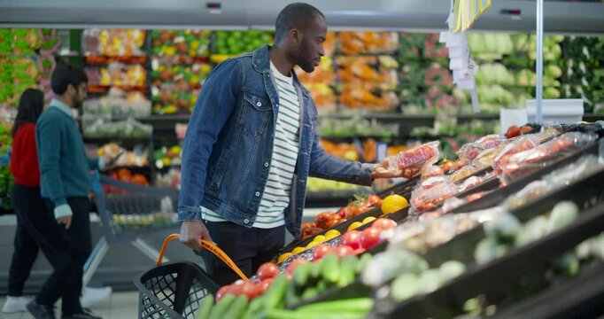 Handsome Black Male Shopper Reaching for Tomatoes on a Vegetable Shelf in the Supermarket, Checking the Quality Before Making His Choice. Young African Man Focused on Eating a Healthy Diet