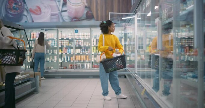 African Female Shopper Opens the Fridge Door in a Supermarket, Reaching for a Milk Bottle and a Dozen Eggs. She Places Them into Her Basket, Ensuring She Gets Fresh Ingredients for Dinner