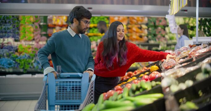 Young Indian Couple Coming to a Grocery Aisle, Carefully Selecting Fresh Ripe Tomatoes and Cucumbers From the Vegetable Shelf. Man and Female Planning to Make a Healthy Salad at Home
