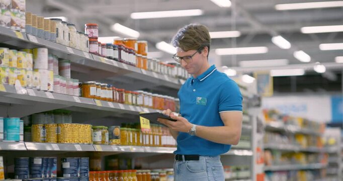 Young Supermarket Manager Using a Tablet Computer to Manage Inventory in a Modern Grocery Store. Caucasian Male Ensuring the Stock is Up to Date and Organized to Satisfy Customers