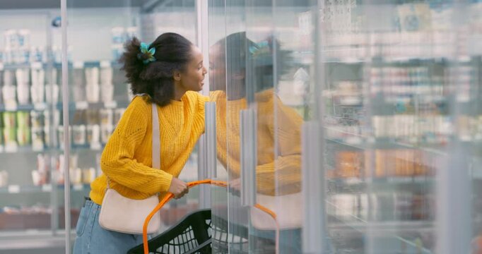 African Female Shopper Opens the Fridge Door in a Supermarket, Reaching for a Milk Bottle and a Dozen Eggs. She Places Them into Her Basket, Ensuring She Gets Fresh Ingredients for Dinner