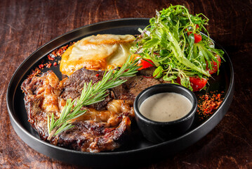 A gourmet steak dish garnished with fresh herbs, served with a side salad and a potato dish on a black plate. The presentation is enhanced with sprinkled seasoning, perfect for restaurant promotions.