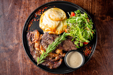 A gourmet steak dish garnished with fresh herbs, served with a side salad and a potato dish on a black plate. The presentation is enhanced with sprinkled seasoning, perfect for restaurant promotions.