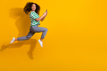 Full body portrait of nice young girl hold phone run jump empty space wear striped t-shirt isolated on yellow color background