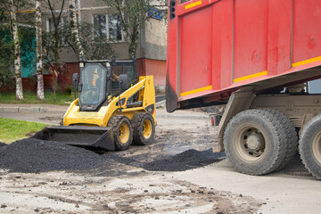 Road works are underway. Hot asphalt is unloaded from the back of a large truck to lay the road. A bulldozer levels the asphalt in an even layer