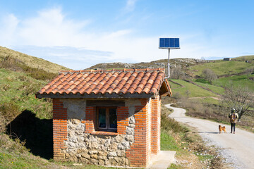 hermit woman and dog walk along a village road in a landscape of countryside and mountains next to a small isolated house with a solar panel and self-sufficient self-consumption energy.