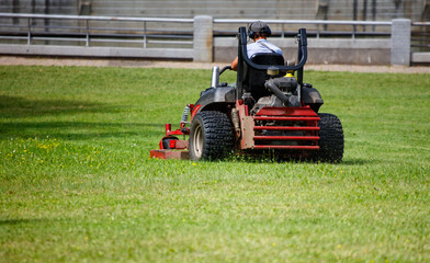 Obraz premium A person operating a lawn mower in a park, cutting the grass. The person is wearing a hat and headphones, focused on the task at hand