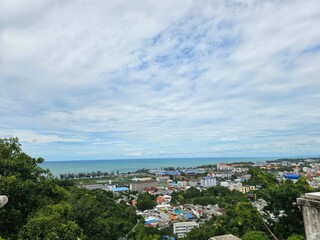 A panoramic view of a coastal town with lush greenery, ocean waves, and a cloudy sky, showcasing the vibrant roofs of houses below.