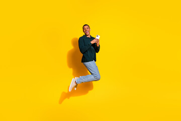 Full size photo of handsome young guy jump hold gadget dressed stylish green garment isolated on yellow color background