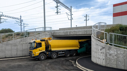 A yellow dump truck gets stuck under a low bridge, showcasing transportation challenges and...