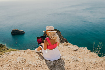 A woman is sitting on a rocky beach with a laptop in front of her. She is wearing a pink top and a straw hat. The beach is near the ocean, and the sky is clear.