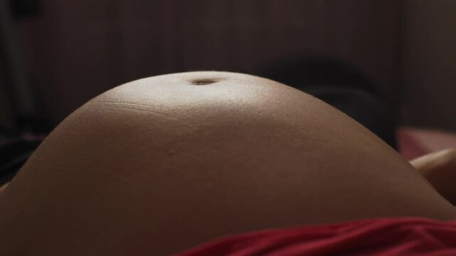 Close-up of pregnant belly showing subtle movements of unborn baby kicking and shifting under stretched skin. Natural lighting highlights texture and shape of belly, creating calm and expectant mood.