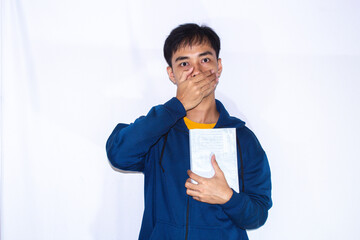 teenager wearing dark blue jacket and yellow shirt, facing forward with shocked expression covering mouth and holding book