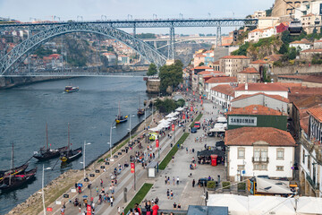 A busy city street in Porto, Portugal, with a bridge in the background. The street is lined with shops and restaurants, and there are many people walking around