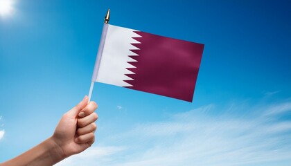 Hand holding Qatar national flag on blue sky.