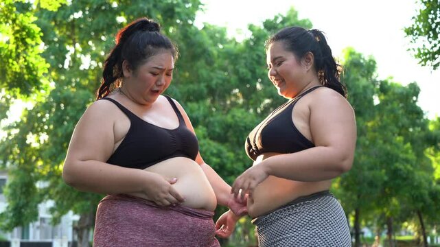 Group of 2 overweight asian woman holding and show belly before exercise in Garden outdoors . two fat female friend warm up for workout outside . Body shaming