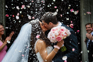 Romantic moment of a newlywed couple kissing while flower petals fall around them during their wedding ceremony, capturing joy and celebration.