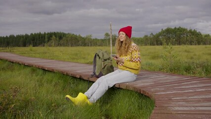 Woman in red beanie and yellow sweater sits on a wooden boardwalk in a grassy meadow, checking her smartphone during a hike. - Powered by Adobe