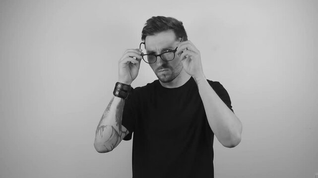 Slow motion footage of a man with tattoos putting on glasses to enhance his vision and squints at the camera because of bad eyesight. Video is filmed in black and white and on a white background.