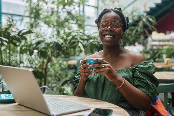 Smiling African American woman holding ceramic cup of tea, she looking at camera while sitting at wooden table