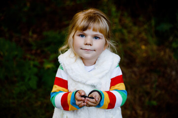 Fototapeta premium Cute little toddler girl making a walk through autumn forest. Happy healthy baby eating blackberries. Sunny warm fall day with child. Active leisure and activity with kids in nature.