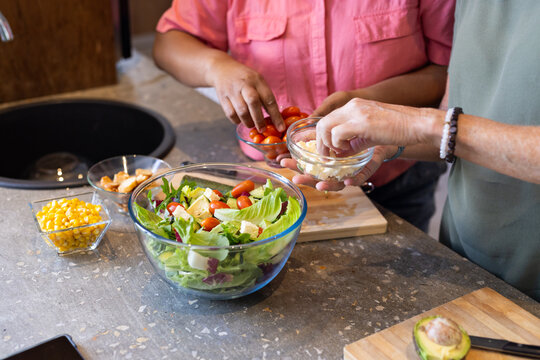 At home, preparing fresh salad, two multiracial female friends adding ingredients in kitchen