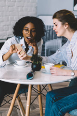 Surprised afro american female blogger feeling shocked from reading text message showed by caucasian friend sitting near, amazed women checking notification for smartphone gadget connected to 4g