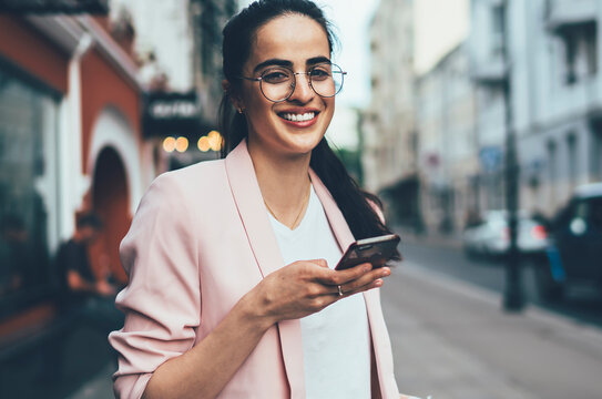 Half length portrait of cheerful Spanish woman in casual outfit enjoying free time for caffee to go, positive millennial woman in spectacles for eyes protection smaling at camera during cell phoning