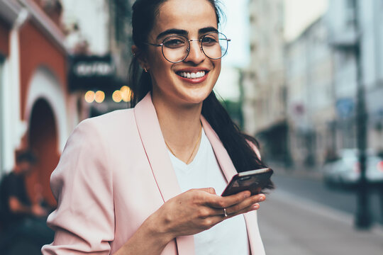Half length portrait of cheerful Spanish woman in casual outfit enjoying free time for caffee to go, positive millennial woman in spectacles for eyes protection smaling at camera during cell phoning
