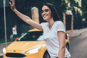 Young charming emotional woman in glasses for better vision gesturing while standing near road for stopping taxi, pretty Georgian hipster girl waving trying catch public transport outdoors