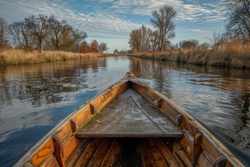 Naklejka premium A serene view from within a wooden boat as it traverses a calm river surrounded by bare trees and golden grassland under a partly cloudy sky
