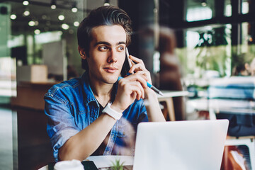 Casual dressed hipster guy talking on smartphone for discussing important information sitting with laptop in coworking space, young man calling on cellular to customer service for consultancy