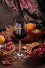 Glass of rich red wine served with fruits on dark wooden background. Autumn still life with wine, grapes and dry leaves in rustic style. Sweater weather concept