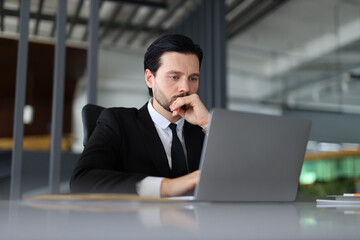 A man in a suit is sitting at a desk with a laptop in front of him. He is deep in thought, possibly working on a project or dealing with a difficult problem. Concept of focus and concentration