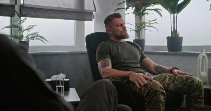 Nervous military man with post traumatic stress disorder sits in chair, listens to psychotherapist during therapy session. Back view of senior psychologist talking to soldier. Over the shoulder.