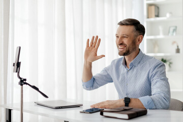 A man is enthusiastically waving while participating in a virtual meeting. He sits at a desk,...