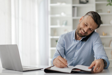 A man sits at a desk in his home office, engaged in a phone conversation while writing notes in a notebook. A laptop is open nearby, indicating a productive work atmosphere.