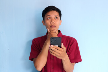 Young Asian man thinking about something while holding mobile phone