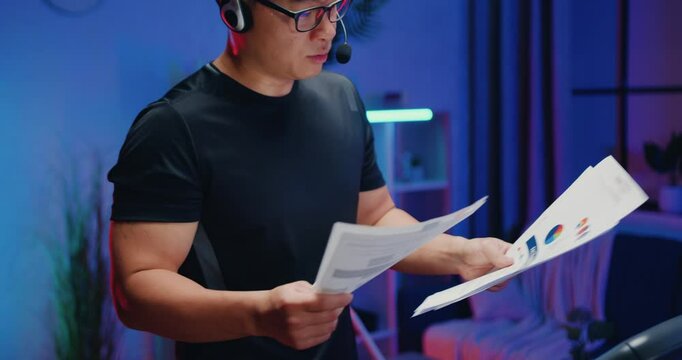 Asian man in headset with mic talking with interlocutor when browsing financial reports with charts durng doing stepping exercises on treadmill at home