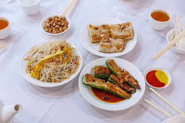 A vibrant spread of Chinese dim sum dishes, including stir-fried noodles, stuffed bell peppers, and radish cake, served with a variety of sauces on a table.