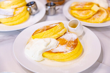 A serving of fluffy souffle pancakes topped with powdered sugar and whipped cream, with a side of syrup, presented beautifully on a white plate