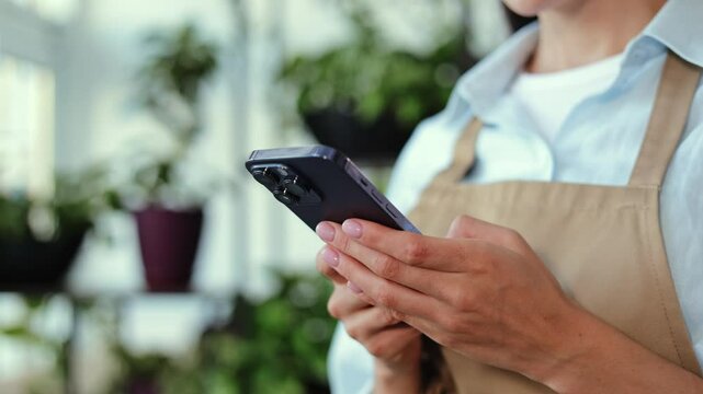 Woman wearing an apron checks her smartphone while working in a plant shop or garden center. Advertisements and social media campaigns showcasing small businesses, plant care or online shopping.