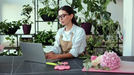 Young woman working as a florist arranges flowers for an order. She is opening her laptop on a desk to check details of the order. There are numerous green plants around the room in terracotta pots.