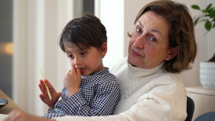 Grandmother sitting with her grandson on her lap at the dining table, watching as he eats. boy is holding a piece of food, fully absorbed in his own thoughts, reflecting a peaceful moment