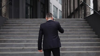 Businessman in a suit walks up set of stairs outside of modern office building while talking on mobile phone. Represent business, success, determination, focus, communication, technology or city life.
