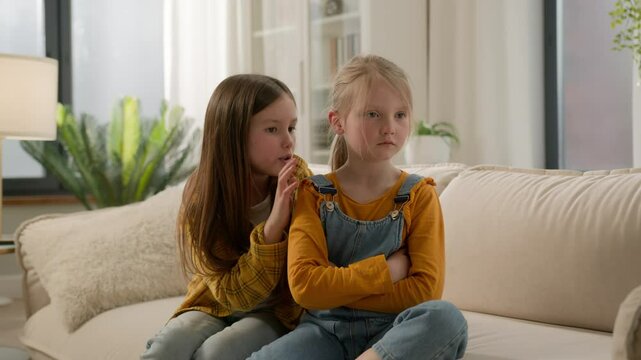 Two little Caucasian kids children siblings daughters girls argument remorseful sister apologizing hugging asking forgiveness younger sister ignoring feel offended angry family quarrel couch indoors
