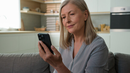 Happy joyful elderly Caucasian woman senior mature old lady use mobile phone smartphone businesswoman typing text message social media chat female 70s granny booking online purchasing living room home