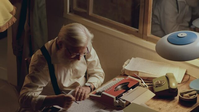 Senior writer sitting at his desk illuminated by warm lamp, crossing out text on his manuscript, crumpling paper and throwing it away as working on book at home. High angle view