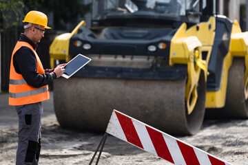 Construction worker using a tablet while overseeing roadwork with a steamroller in the background