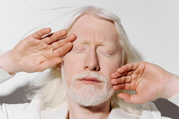 Portrait of a serene person with pale skin and long white hair, gently closing their eyes, hands framing their face against a minimalistic white background